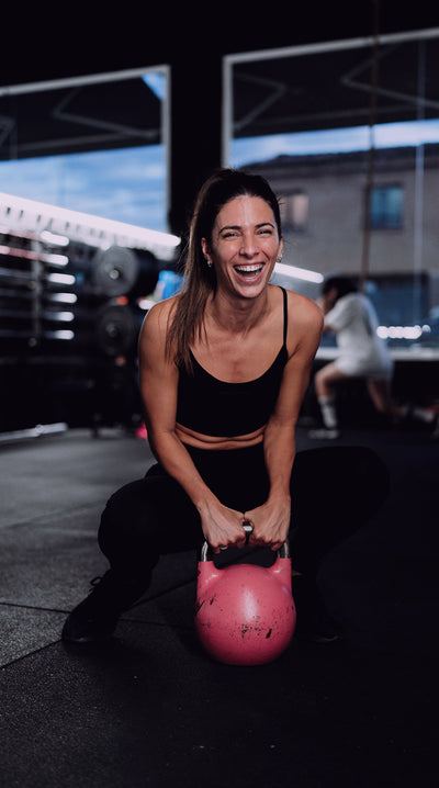 Mujer sonriendo en cuclillas en un box de crosstraining con una kettlebell rosa 